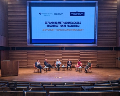 Six adults dressed in business casual attire are seated in a row on stage with a large screen above their heads that says Expanding Methadone Access in Correctional Facilities.