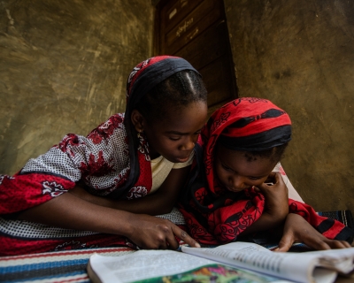 Two girls dressed in red and black garments sit on the floor reading USAID malaria educational materials in Zanzibar, Tanzania. 