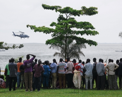 Dozens of bystanders look on as they watch rescuers in a helicopter search the site of a capsized cruise boat on Lake Victoria near Mutima village, south of Kampala, Uganda, on a grey, cloudy day.