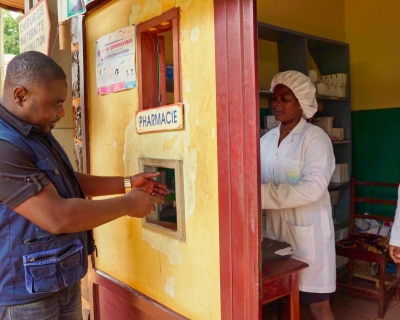 A man wearing a blue vest puts his hands out to receive medicine through a window cut out in a yellow-painted pharmacy.