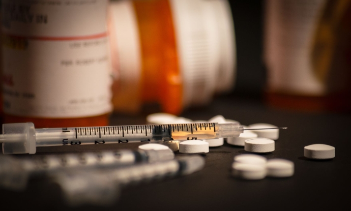 White pills are scattered on the counter near a few orange pill bottoms. A syringe is next to the scattered pills.